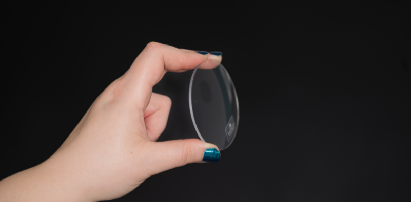 A hand holding a spectacle lens between their fingers on a black background.