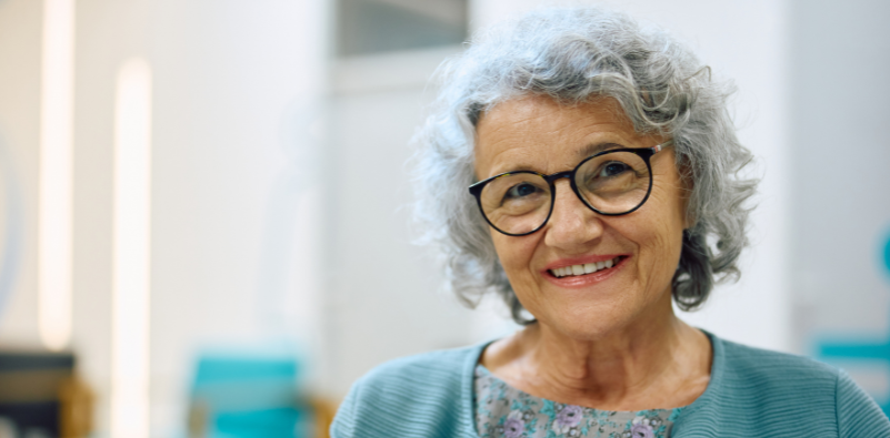 A senior lady wearing black glasses smiling at the camera.